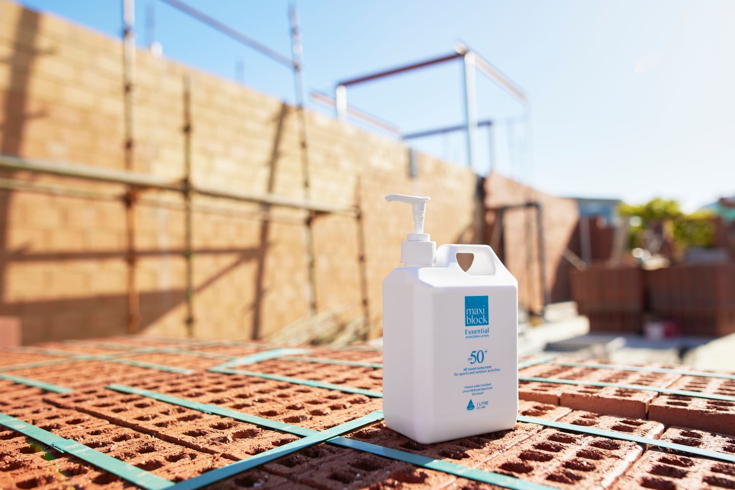 Maxiblock Essential SPF 50+ 1 litre pump bottle on a brick construction site with scaffolding in background