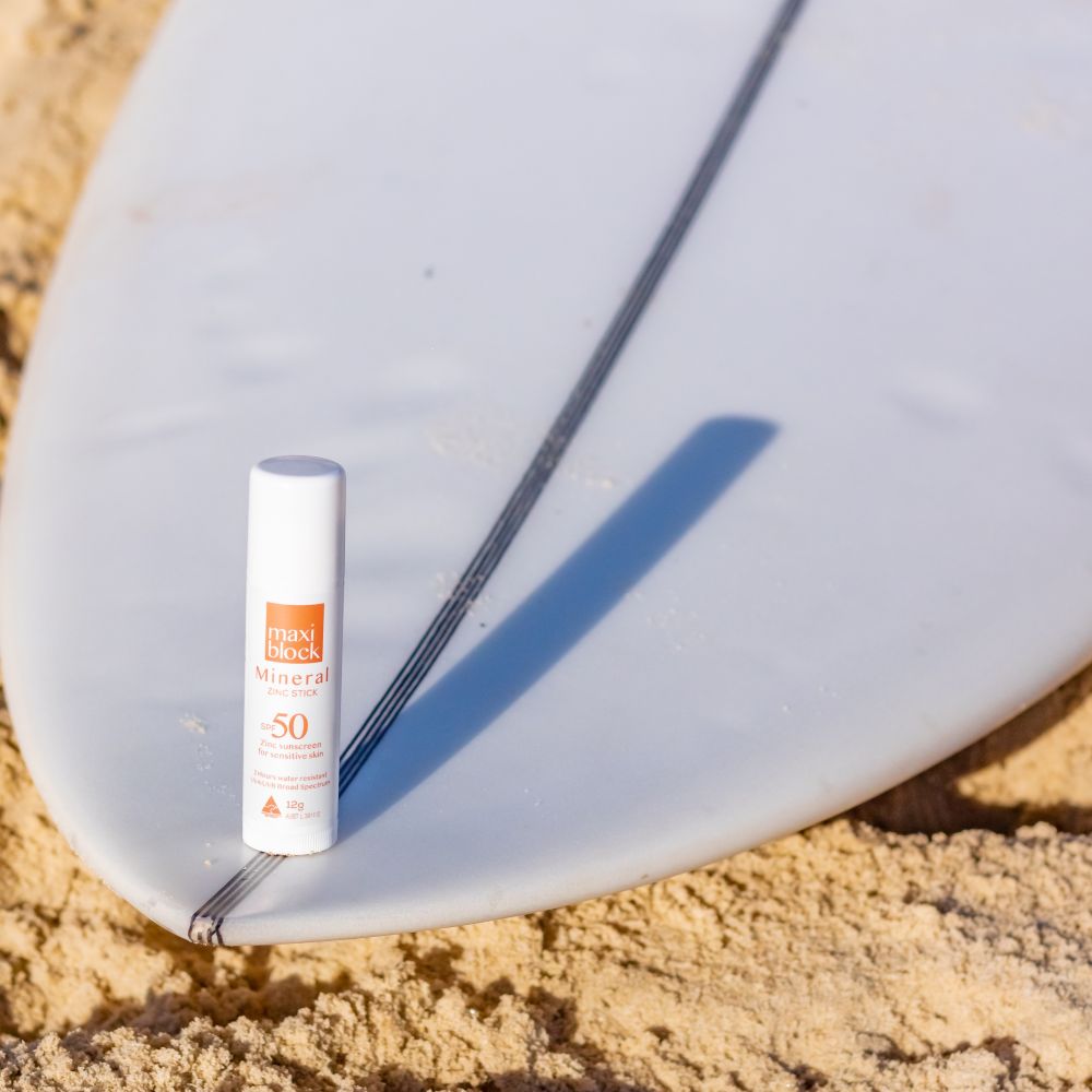 Maxiblock Mineral Zinc Stick Clear SPF 50+ leaning against a surfboard on the sand
